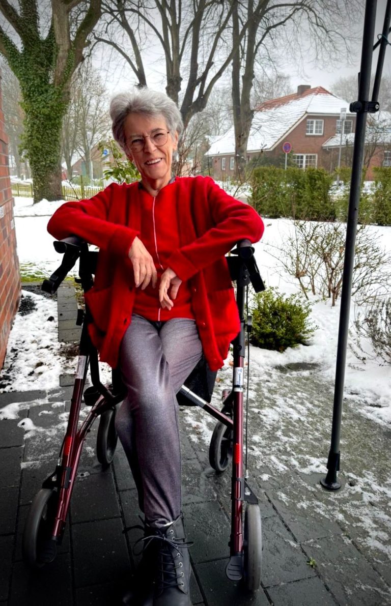 Eine ältere Frau in einem Rollstuhl sitzt im Schnee vor einem Haus.