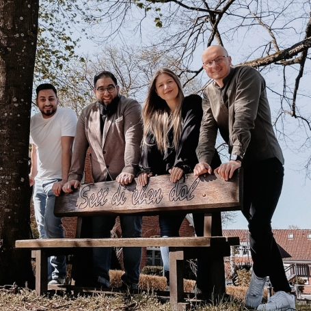 Vier Personen lehnen lässig an einer Bank unter einem Baum im Freien. Plattdeutsch