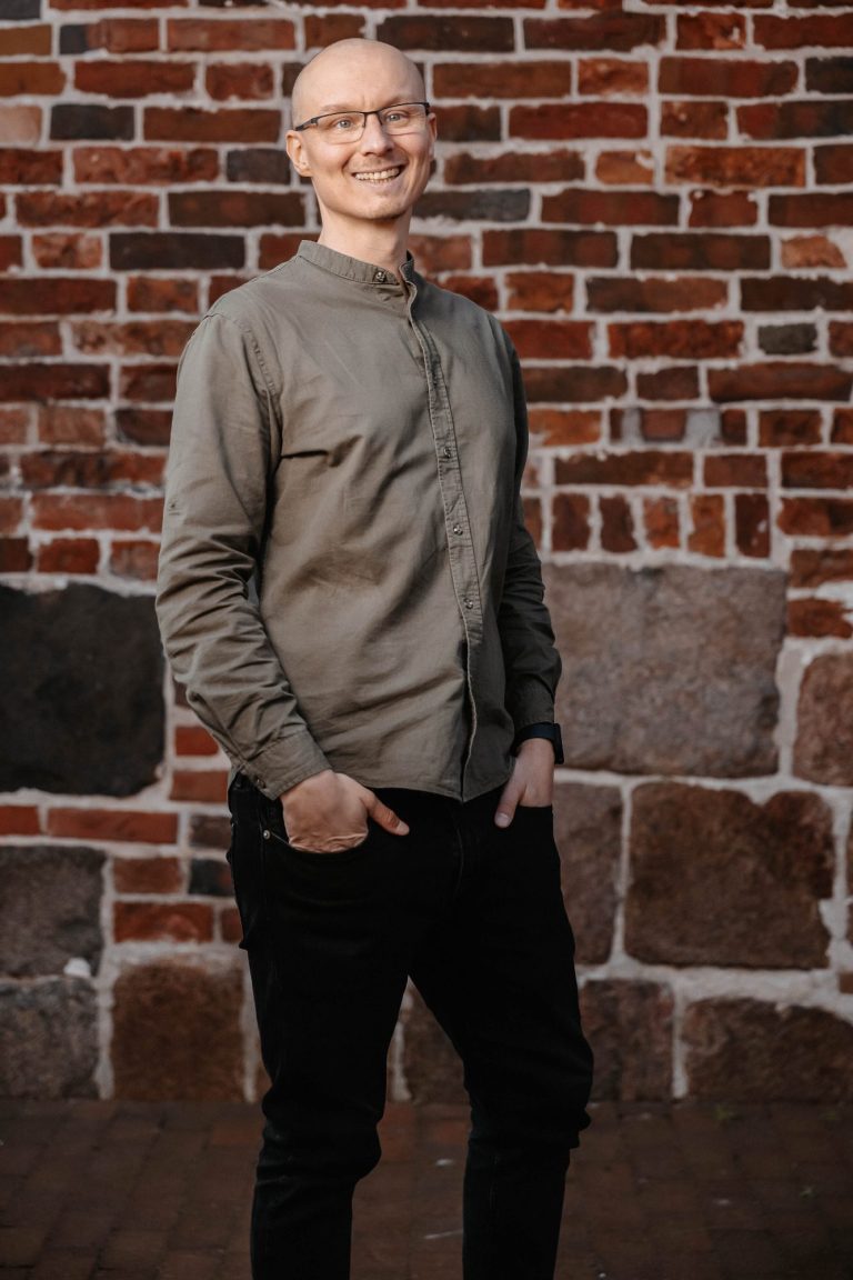 Pflegedienst Ostfriesland Mann mit Brille, lächelnd, steht vor einer roten Backsteinwand.